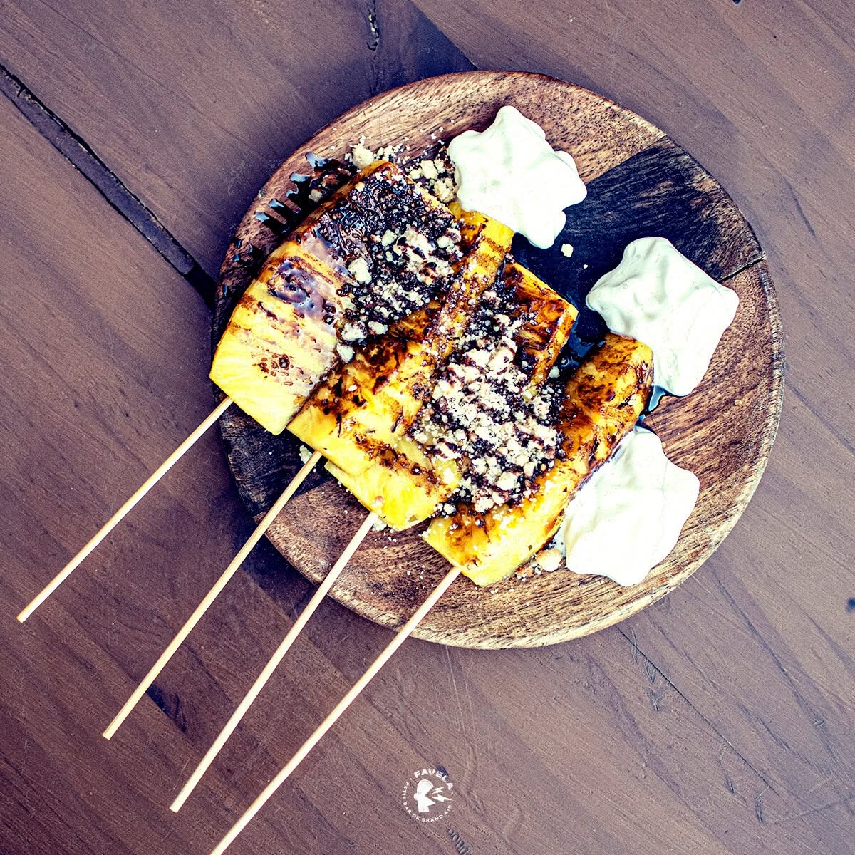 Brochettes d'ananas grillé au chocolat et crème sur plat en bois. Logo Favela.