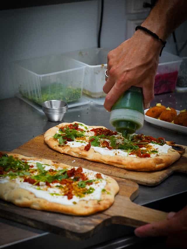 Chef garnissant une focaccia burrata, tomates séchées et roquette avec de l'huile aux herbes.