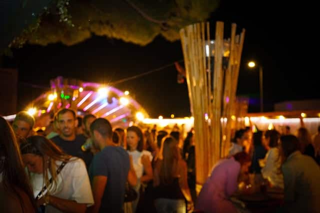 Foule à une soirée festive en plein air sous des lumières colorées et structures en bois.